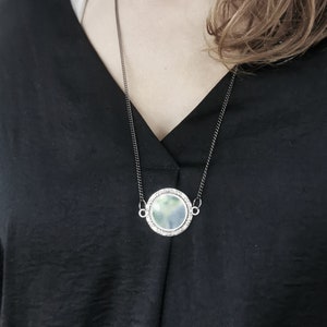 K&ouml;nnte beinhalten: Eine silberne Kettenkette mit einem runden Anh&auml;nger mit einem wolkigen, gr&uuml;n-blauen Design.