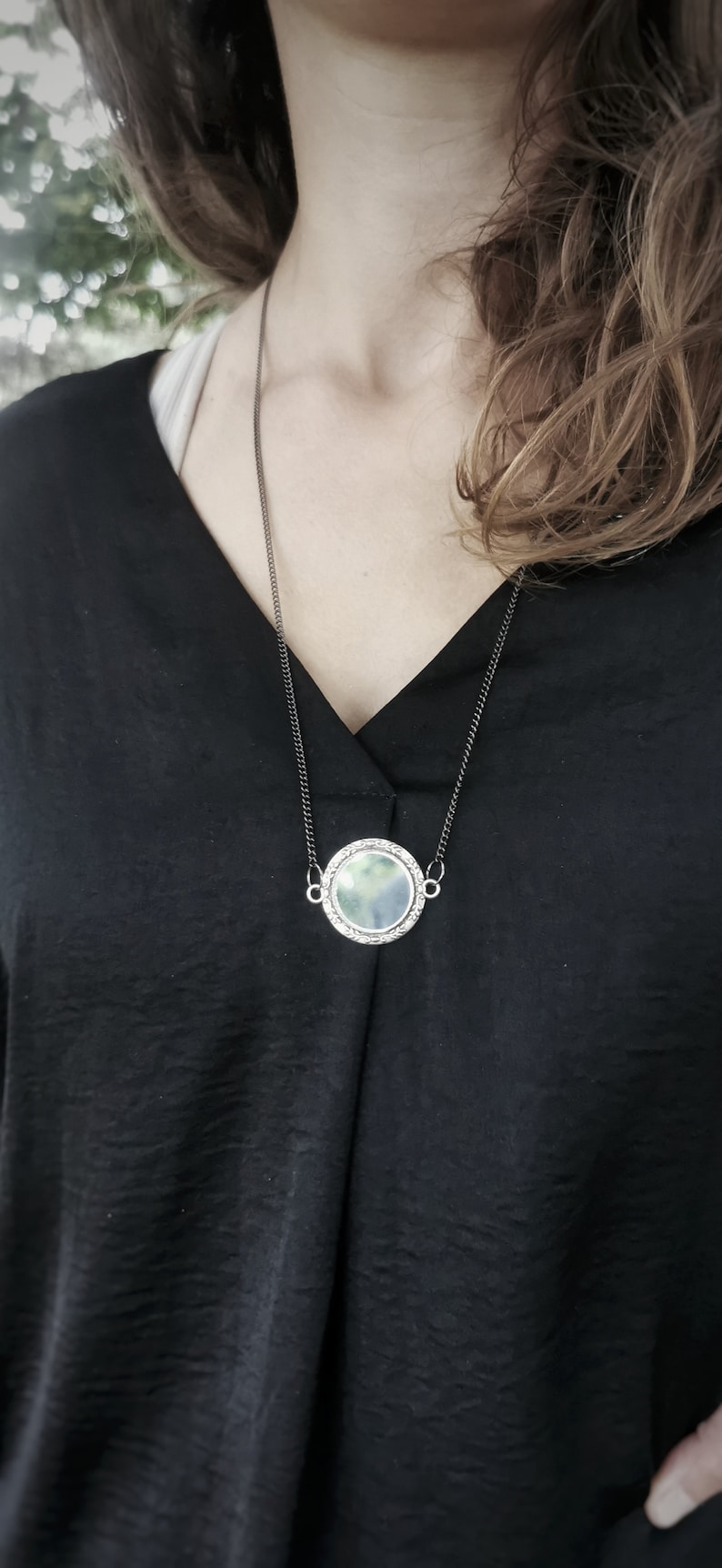 K&ouml;nnte beinhalten: Eine silberne Kettenkette mit einem runden Anh&auml;nger mit einem wolkigen, gr&uuml;n-blauen Design.