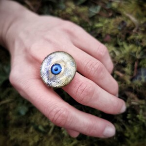 以下が含まれることがあります： 青い目のデザインが施された金の指輪。指につけられており、背景はぼやけている。