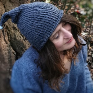 Puede incluir: Un gorro azul de punto con la parte superior puntiaguda, usado por una persona con el pelo largo y castaño. El gorro tiene una textura de punto de canalé y un diseño sencillo.