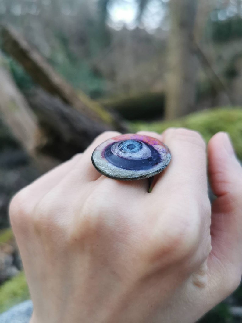 May include: A ring with a large, round, eye-shaped design. The eye is black with a white center and a blue iris. The ring is made of glass and has a silver band.