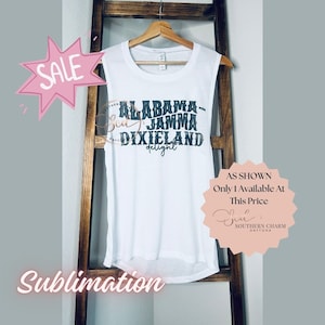 May include: White sleeveless shirt with black text that reads "Alabama Jamm DixieLand delight". The shirt is on a wooden hanger and has a pink starburst with the word "Sale" on it.
