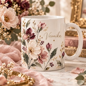 May include: A white ceramic mug featuring a floral design with shades of pink, burgundy, and green, accented with gold details. The name Veronika is printed in a gold script font on the front, accompanied by a small gold heart symbol.