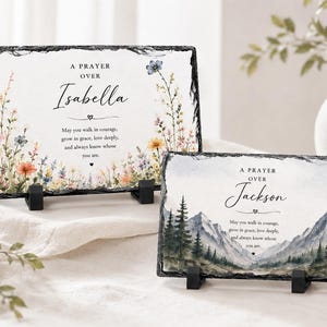 May include: Two rectangular slate plaques with rough, dark edges displayed on black stands. The larger plaque features a floral design and the text A PRAYER OVER Isabella. The smaller plaque shows a mountain landscape and the text A PRAYER OVER Jackson.