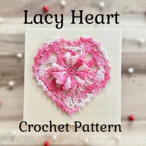 Puede incluir: Un patrón de corazón de ganchillo calado, en tonos rosa y blanco, adornado con una flor central, sobre un fondo crema. El corazón presenta un diseño detallado y calado. El texto "Lacy Heart Crochet Pattern" se muestra en negro.