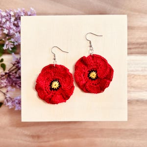 May include: A pair of handmade red crochet poppy earrings with silver-colored hooks. The center of each flower is yellow with black detailing. The earrings are displayed on a light wooden surface.