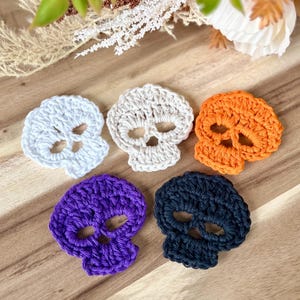 May include: Five crocheted skull shapes in white, beige, orange, purple, and navy blue. The skulls are handmade with visible stitch detail. The skulls are arranged on a wooden surface, perfect for Halloween or spooky decor.