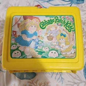 May include: Yellow plastic lunchbox with a Cabbage Patch Kids design. The image features a Cabbage Patch Kid doll watering a garden of cabbage patch kids. The text on the lunchbox reads "Cabbage Patch Kids".
