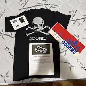 May include: Black t-shirt with a white skull and crossbones graphic and the word "GOONIES". Accompanied by a certificate of authenticity, a patch, and a Warner Bros. feature card.