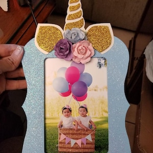 May include: A light blue picture frame with a unicorn design. The unicorn has a gold glitter horn and ears, and is decorated with pink, purple, and blue flowers. The frame has a glitter finish and is perfect for displaying a photo of twins.