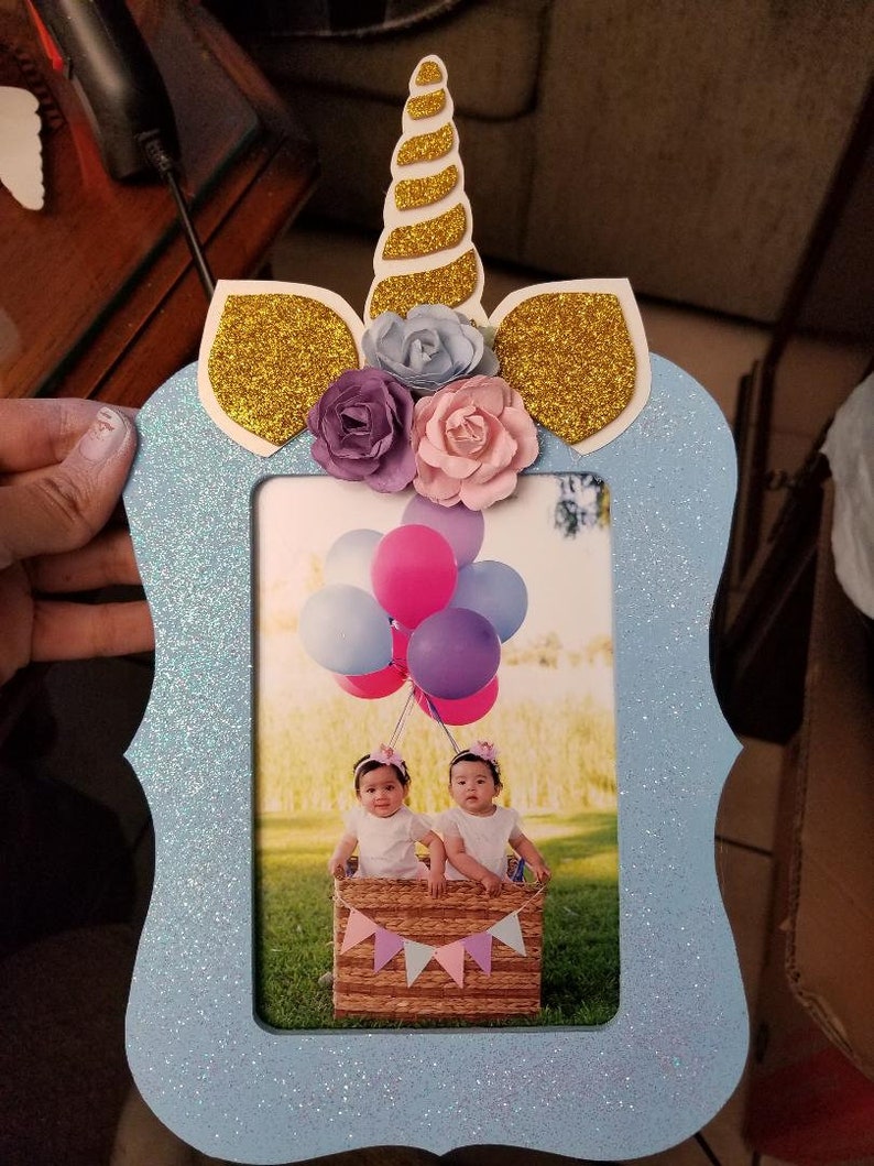 May include: A light blue picture frame with a unicorn design. The unicorn has a gold glitter horn and ears, and is decorated with pink, purple, and blue flowers. The frame has a glitter finish and is perfect for displaying a photo of twins.
