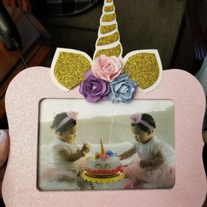 May include: Pink glitter unicorn picture frame with a gold glitter horn and ears. The frame has a photo of two young girls in matching dresses and headbands, celebrating a birthday with a unicorn cake.