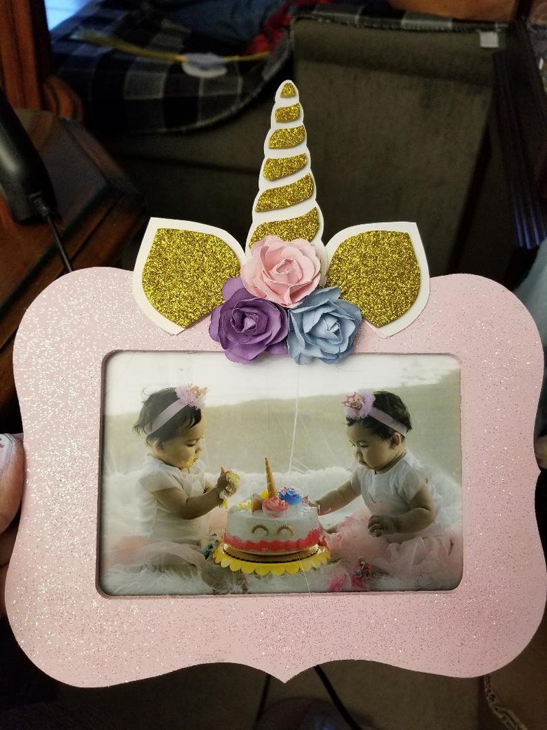 May include: Pink glitter unicorn picture frame with a gold glitter horn and ears. The frame has a photo of two young girls in matching dresses and headbands, celebrating a birthday with a unicorn cake.