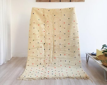 May include: A cream-coloured Moroccan rug with a scattered pattern of small, colourful dots in red, blue, and green. The rug has a fringe border and is folded in half.