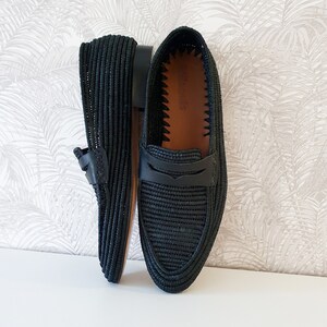 Puede incluir: Un par de mocasines de cuero negro tejido con un detalle de correa de cuero. Los mocasines tienen un forro de cuero marr&oacute;n y una textura tejida.