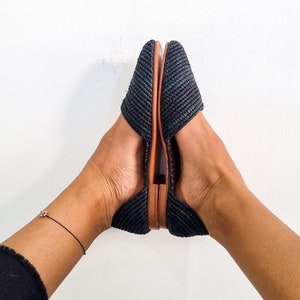 May include: A pair of black woven flat shoes with brown leather soles. The shoes are worn by a person with bare feet.