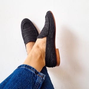 May include: A pair of black woven slip-on shoes with a brown leather sole. The shoes are worn by a person wearing blue jeans.