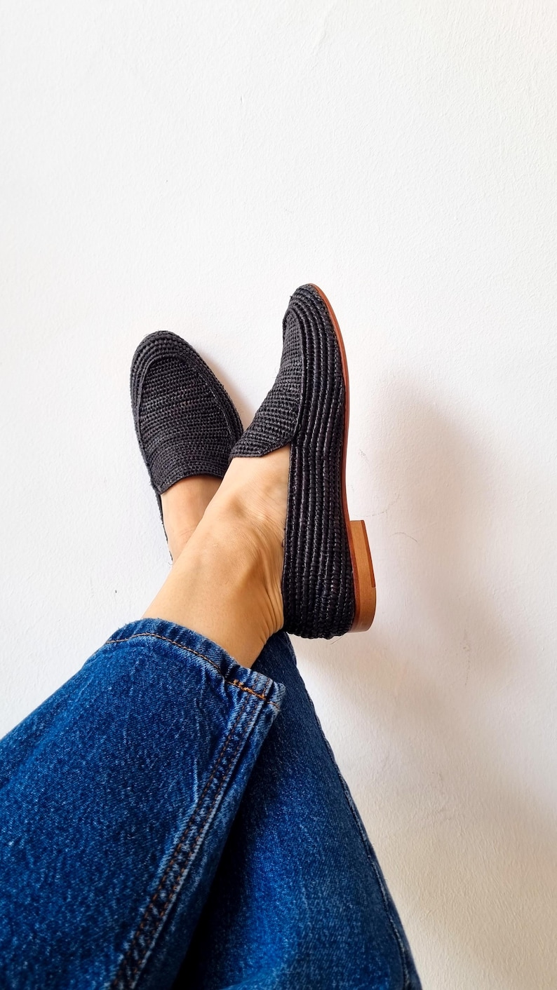 May include: A pair of black woven slip-on shoes with a brown leather sole. The shoes are worn by a person wearing blue jeans.