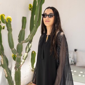 May include: A woman wearing a black dress with sheer striped sleeves and black sunglasses. She is standing in front of a cactus plant.