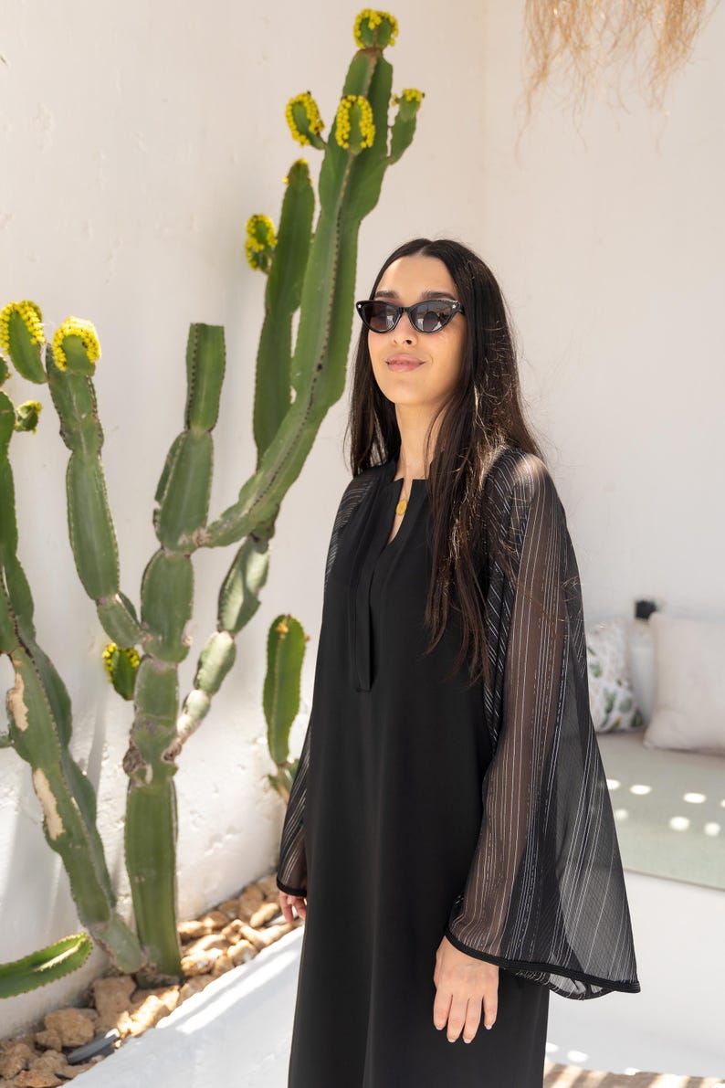 May include: A woman wearing a black dress with sheer striped sleeves and black sunglasses. She is standing in front of a cactus plant.