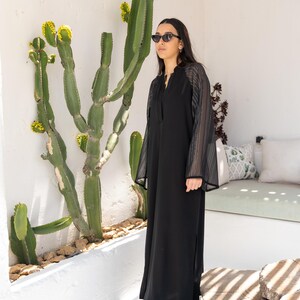 May include: A black maxi dress with sheer, striped sleeves. The dress has a V-neckline and a side slit. The model is wearing black sunglasses and standing in front of a cactus.