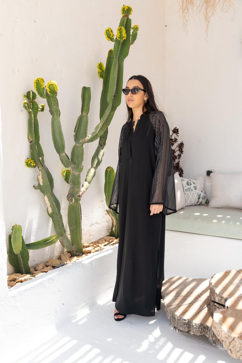 May include: A black maxi dress with sheer, striped sleeves. The dress has a V-neckline and a side slit. The model is wearing black sunglasses and standing in front of a cactus.