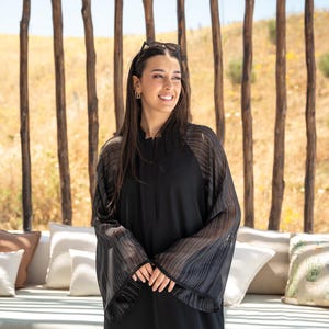 May include: A woman wearing a black abaya with sheer, striped sleeves. She is standing in front of a wooden slat wall and smiling.