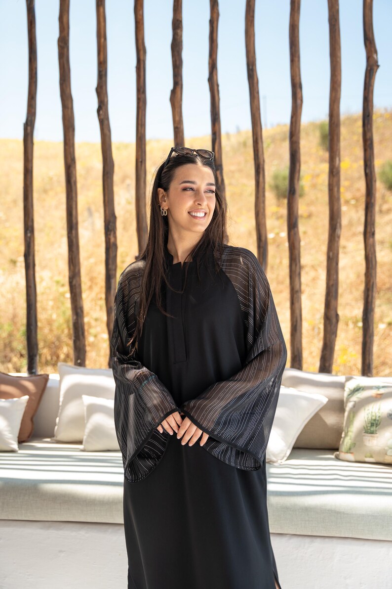 May include: A woman wearing a black abaya with sheer, striped sleeves. She is standing in front of a wooden slat wall and smiling.