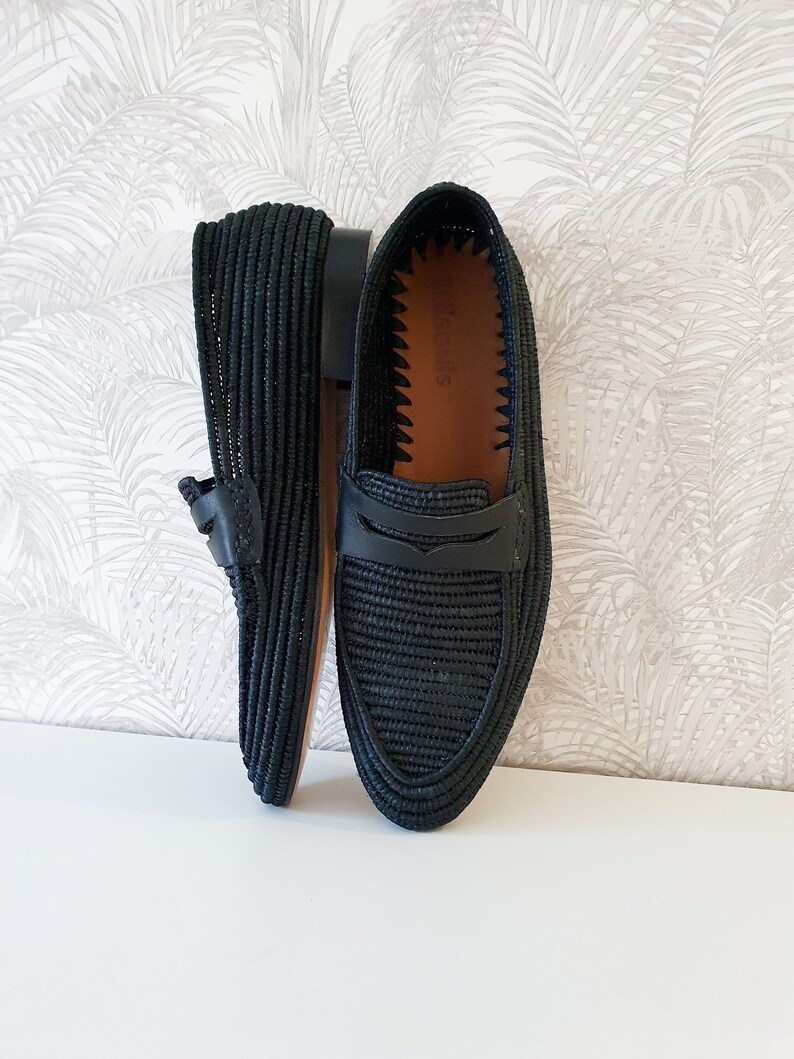 Puede incluir: Un par de mocasines de cuero negro tejido con un detalle de correa de cuero. Los mocasines tienen un forro de cuero marr&oacute;n y una textura tejida.