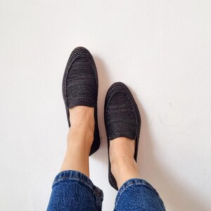 May include: A pair of black woven slip-on shoes with a person wearing blue jeans.