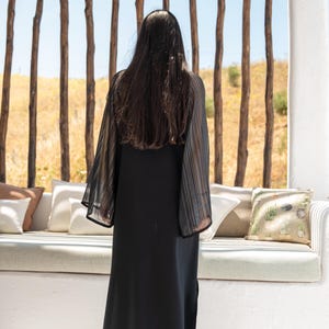 May include: A woman wearing a black maxi dress with sheer, striped sleeves. She is standing in front of a white wall with a wooden slat backdrop.