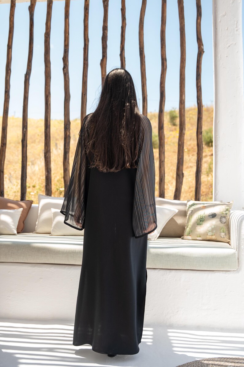May include: A woman wearing a black maxi dress with sheer, striped sleeves. She is standing in front of a white wall with a wooden slat backdrop.