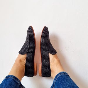 May include: A pair of black woven loafers with brown leather soles. The loafers are worn by a person wearing blue jeans.