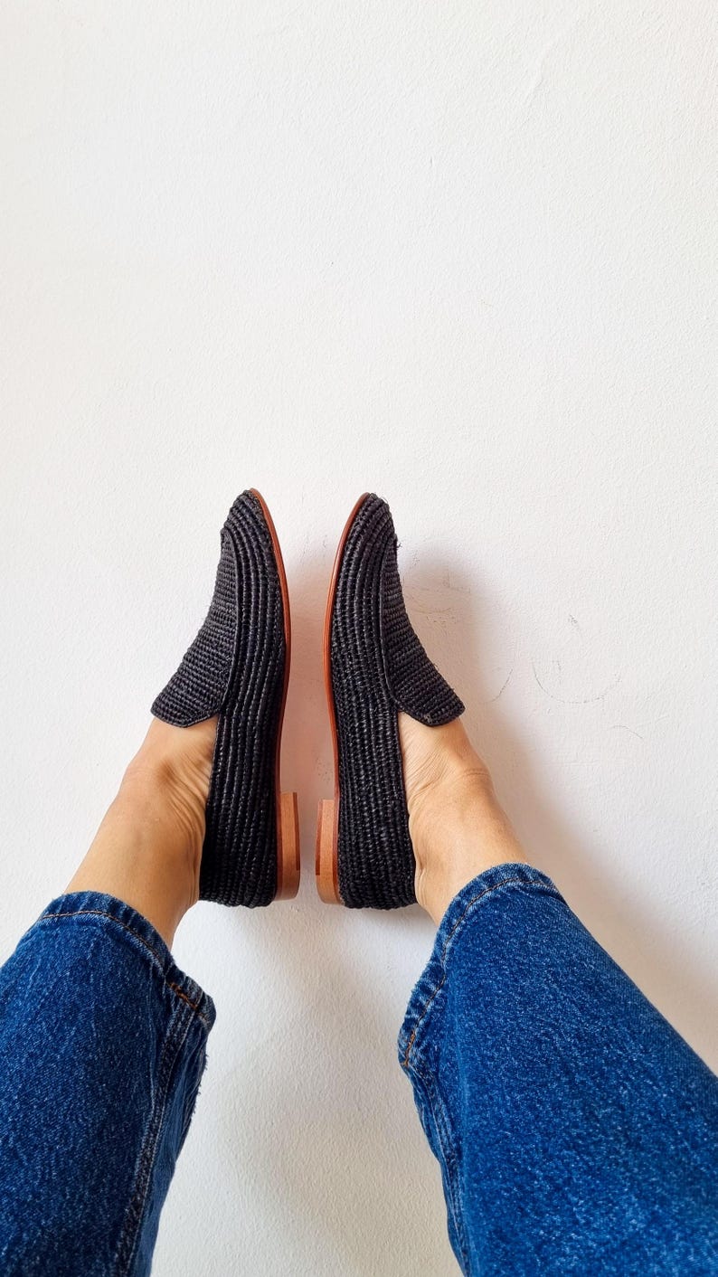 May include: A pair of black woven loafers with brown leather soles. The loafers are worn by a person wearing blue jeans.