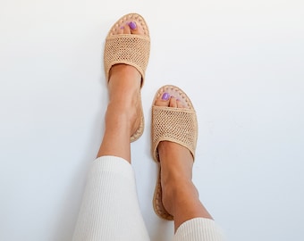 May include: A pair of beige woven mesh slide sandals with a light brown sole. The sandals are worn on a person's feet, with the person's legs and white pants visible.