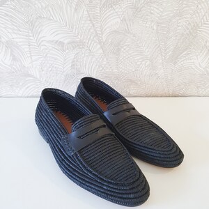 Puede incluir: Un par de mocasines negros de cuero tejido con una tira de cuero en la parte superior. Los mocasines tienen una punta puntiaguda y una suela plana.