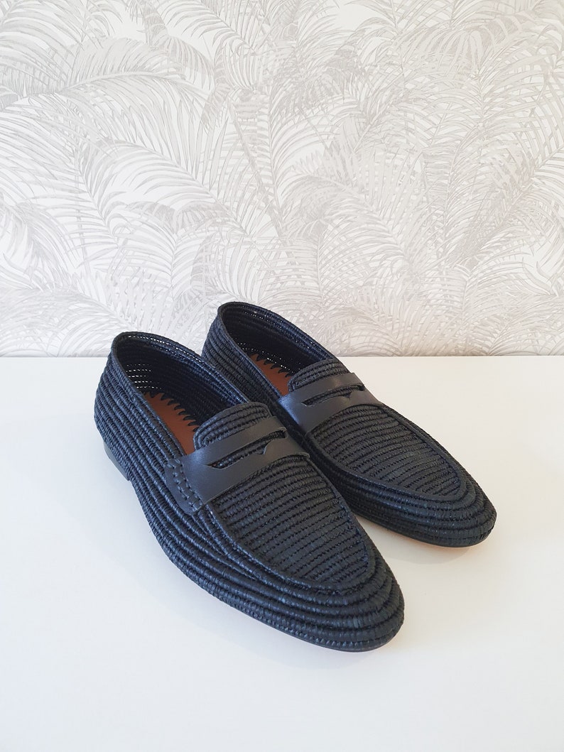 Puede incluir: Un par de mocasines negros de cuero tejido con una tira de cuero en la parte superior. Los mocasines tienen una punta puntiaguda y una suela plana.