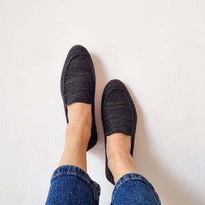 May include: A pair of black woven slip-on shoes with a person wearing blue jeans.