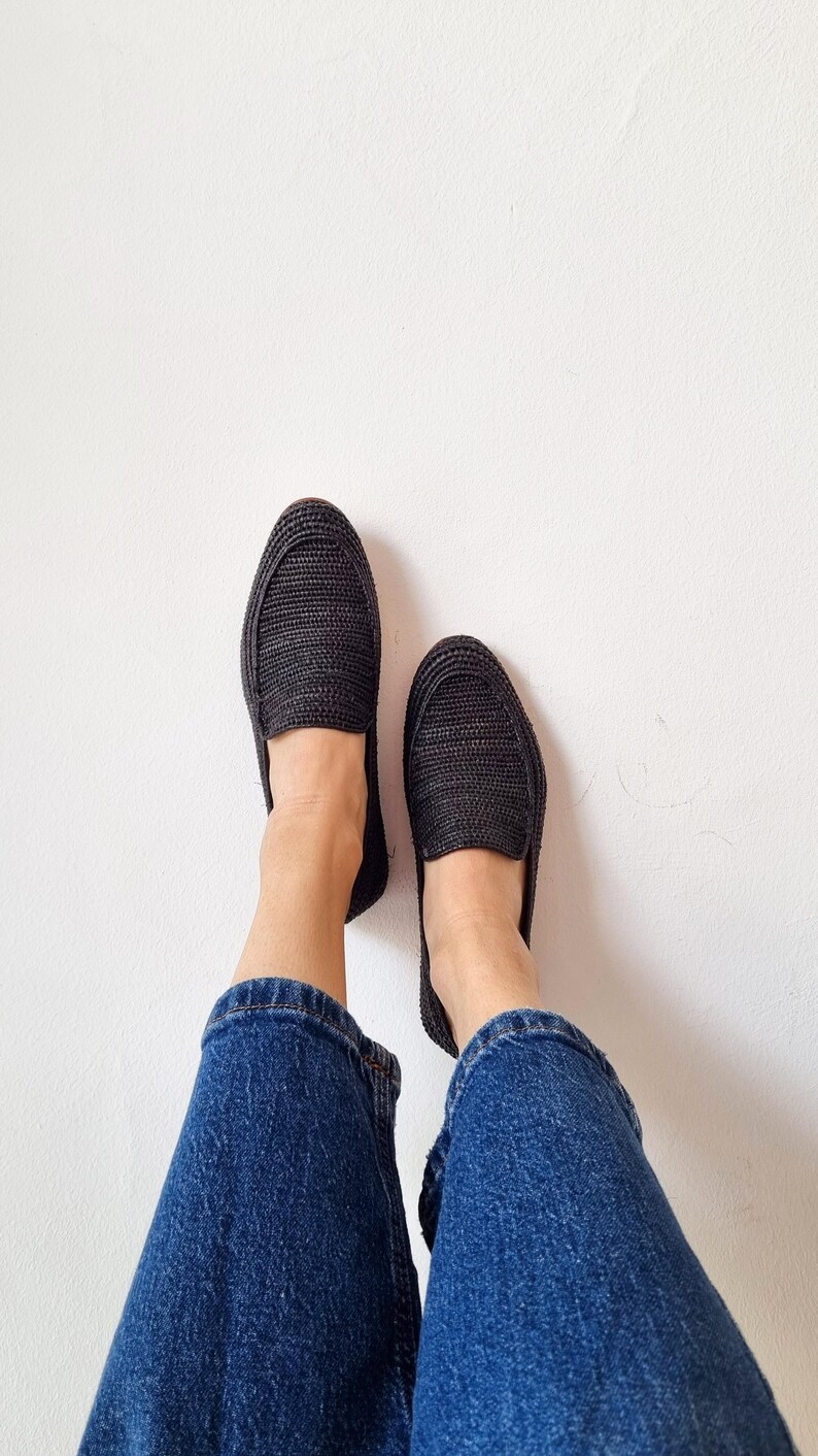 May include: A pair of black woven slip-on shoes with a person wearing blue jeans.