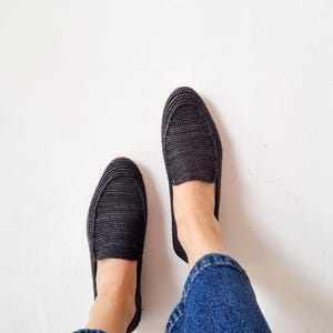 May include: A pair of black woven slip-on shoes with a person wearing blue jeans.