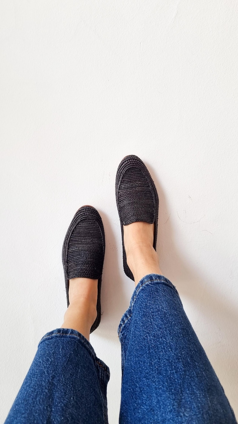 May include: A pair of black woven slip-on shoes with a person wearing blue jeans.