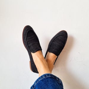 May include: A pair of black woven slip-on shoes with a brown sole. The shoes are worn by a person wearing blue jeans.