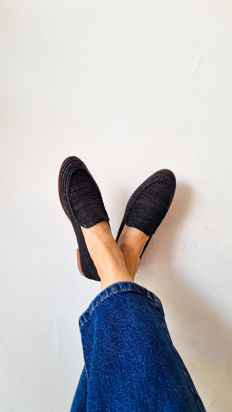 May include: A pair of black woven slip-on shoes with a brown sole. The shoes are worn by a person wearing blue jeans.