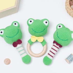 Op de afbeelding: Drie gehaakte groene kikker speelgoedjes. Twee van de kikkers zijn bevestigd aan houten rammelaars, een met een rood-wit gestreepte handvat en de andere met een groen-wit gestreepte handvat. De derde kikker is bevestigd aan een houten ring.