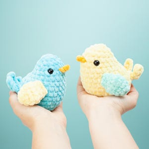 Puede incluir: Dos pájaros de peluche hechos a mano, uno azul claro y otro amarillo, con ojos negros y picos naranjas. El pájaro azul tiene un ala amarilla, y el pájaro amarillo tiene un ala azul claro. Los pájaros son suaves.