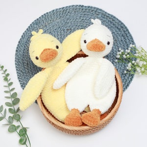 May include: Two crocheted duck plushies, one yellow and one white, sitting in a wicker basket. The ducks have brown beaks and orange feet.