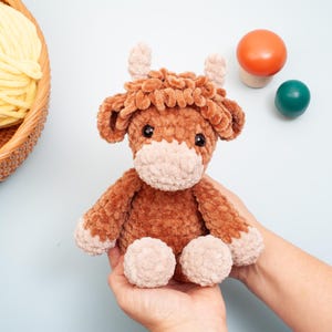 May include: A handmade brown and beige crochet cow plush toy with black safety eyes. The plush cow has a fluffy body, head, and legs. The cow is being held in a person's hands. A basket of yellow yarn is in the background.