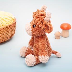 May include: A plush brown and tan Highland cow stuffed animal with a fluffy texture. The cow has small horns, a tan face, and is sitting on a light blue surface. A basket of yellow yarn and wooden mushrooms are in the background.