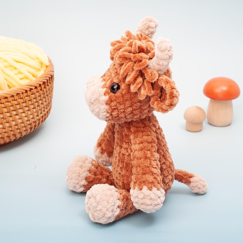 May include: A plush brown and tan Highland cow stuffed animal with a fluffy texture. The cow has small horns, a tan face, and is sitting on a light blue surface. A basket of yellow yarn and wooden mushrooms are in the background.