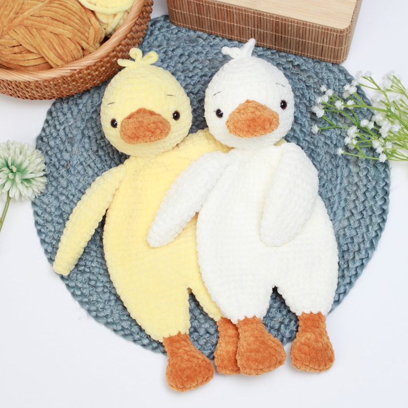 May include: Two crocheted plush duck toys, one yellow and one white, with orange beaks and feet. The toys are sitting on a blue and gray textured surface.